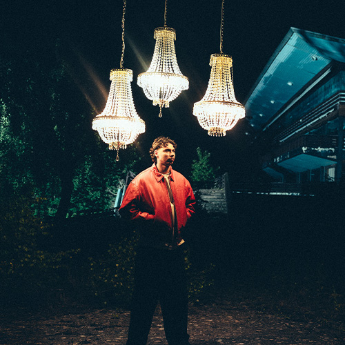 photo du rappeur GEORGIO, mains dans les poches, avec une veste rouge. On le voit debout, dehors. Au-dessus de lui pendent trois lustres.