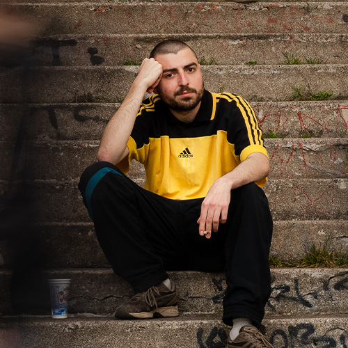 Photo du rappeur lyonnais OKIS, assis sur une scalier, fixant l'objectif. On voit autour des ombres de personnes.